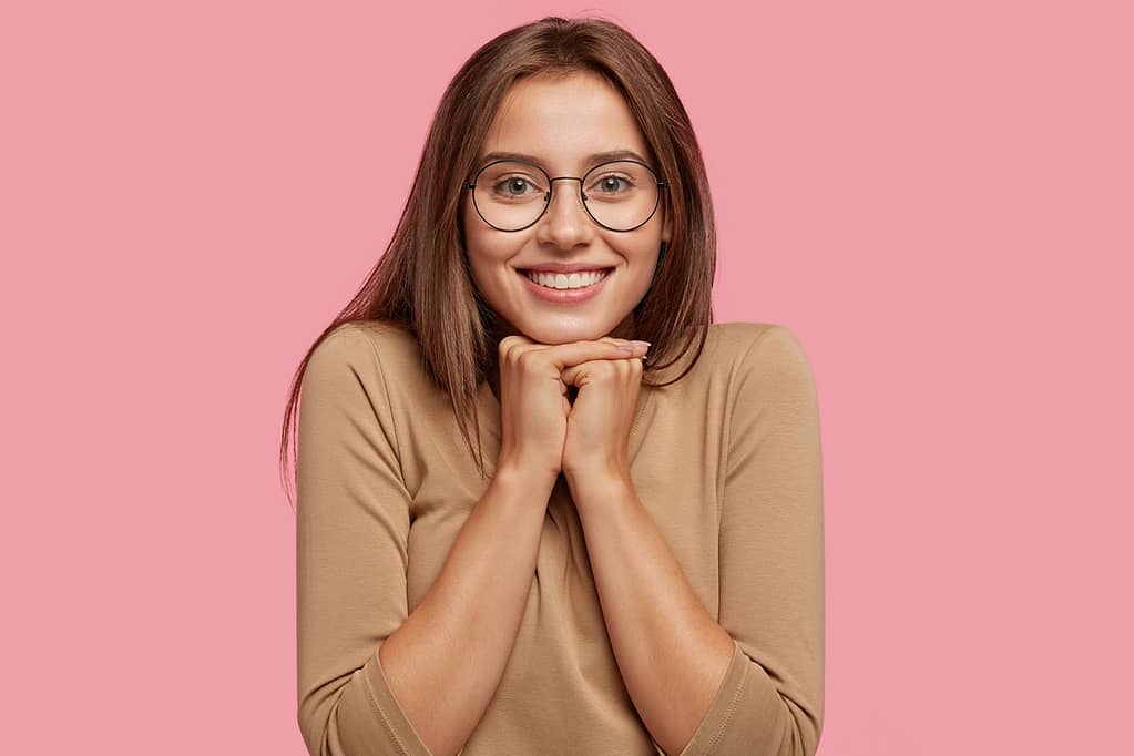 The Wonders of Arm Liposuction 1 – arm liposuction The Wonders Arm Liposuction, Photo of pleasant looking brunette Caucasian woman has toothy smile, holds chin with both hands, dressed in beige casual clothes, poses over pink studio background. Positive emotions concept