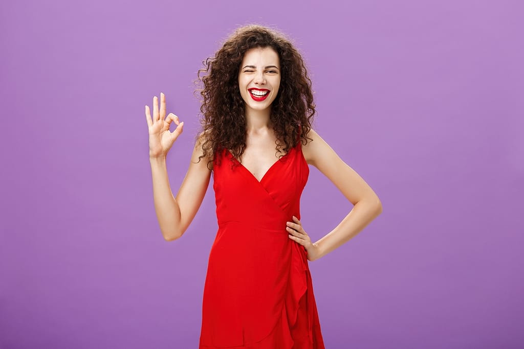 I got you covered. Happy outgoing good-looking elegant european female with curly hairstyle in red fashionable dress, make-up holding hand on waist showing okay sign in approval and smiling assuring.