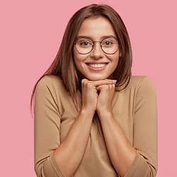 The Wonders Arm Liposuction, Photo of pleasant looking brunette Caucasian woman has toothy smile, holds chin with both hands, dressed in beige casual clothes, poses over pink studio background. Positive emotions concept