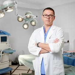 Arm Liposuction Cost Front view of man surgeon in glasses looking at camera with serious expression while standing in operating room. Handsome male doctor with crossed arms wearing white lab coat. Concept of medicine